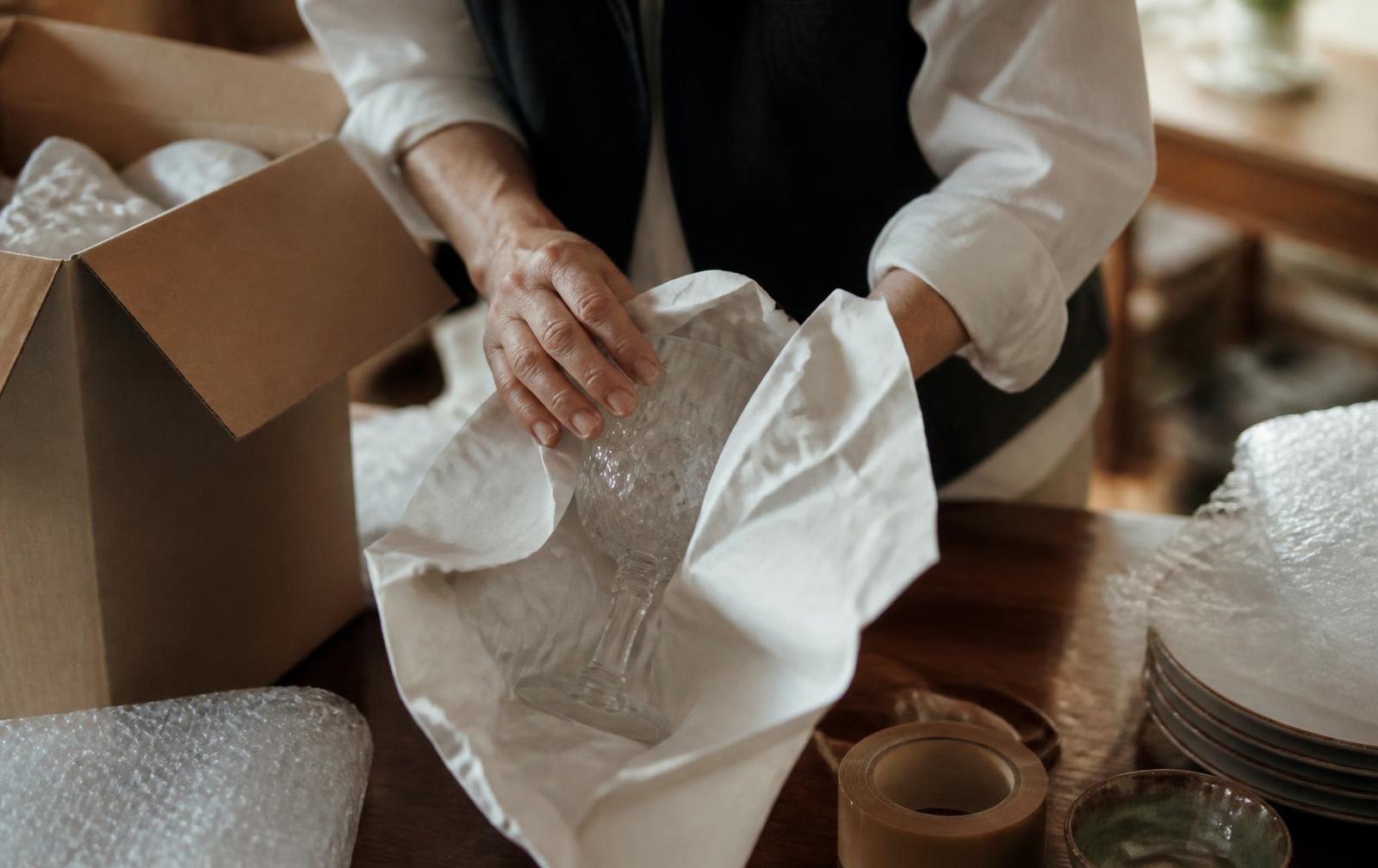 Glassware being carefully wrapped in packing paper beside a moving carton